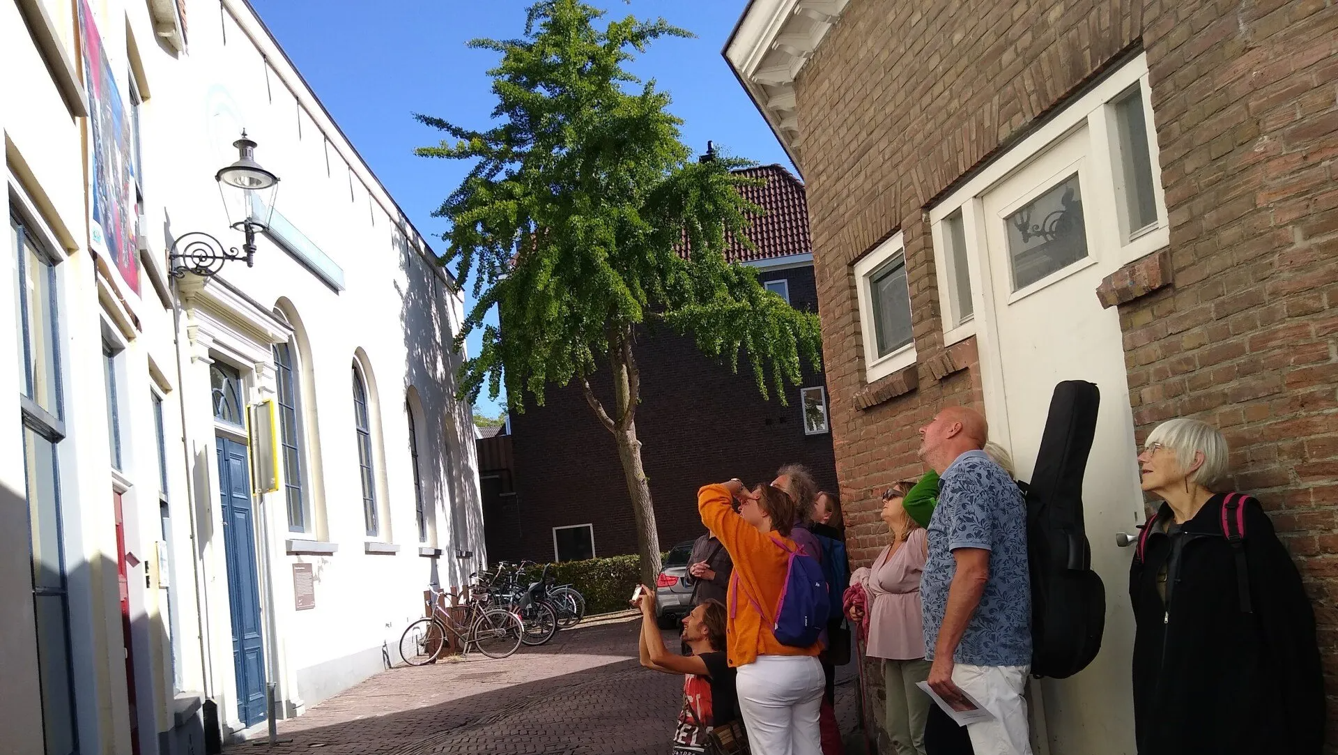 18 september 2020: Kunstenaars en belangstellenden bekijken de kunst voor de vrede bij het Etty Hillesum Centrum. Een groep mensen staat op een geplaveide straat en kijkt omhoog naar de gevel van een gebouw in een zonnig, historisch stadje.