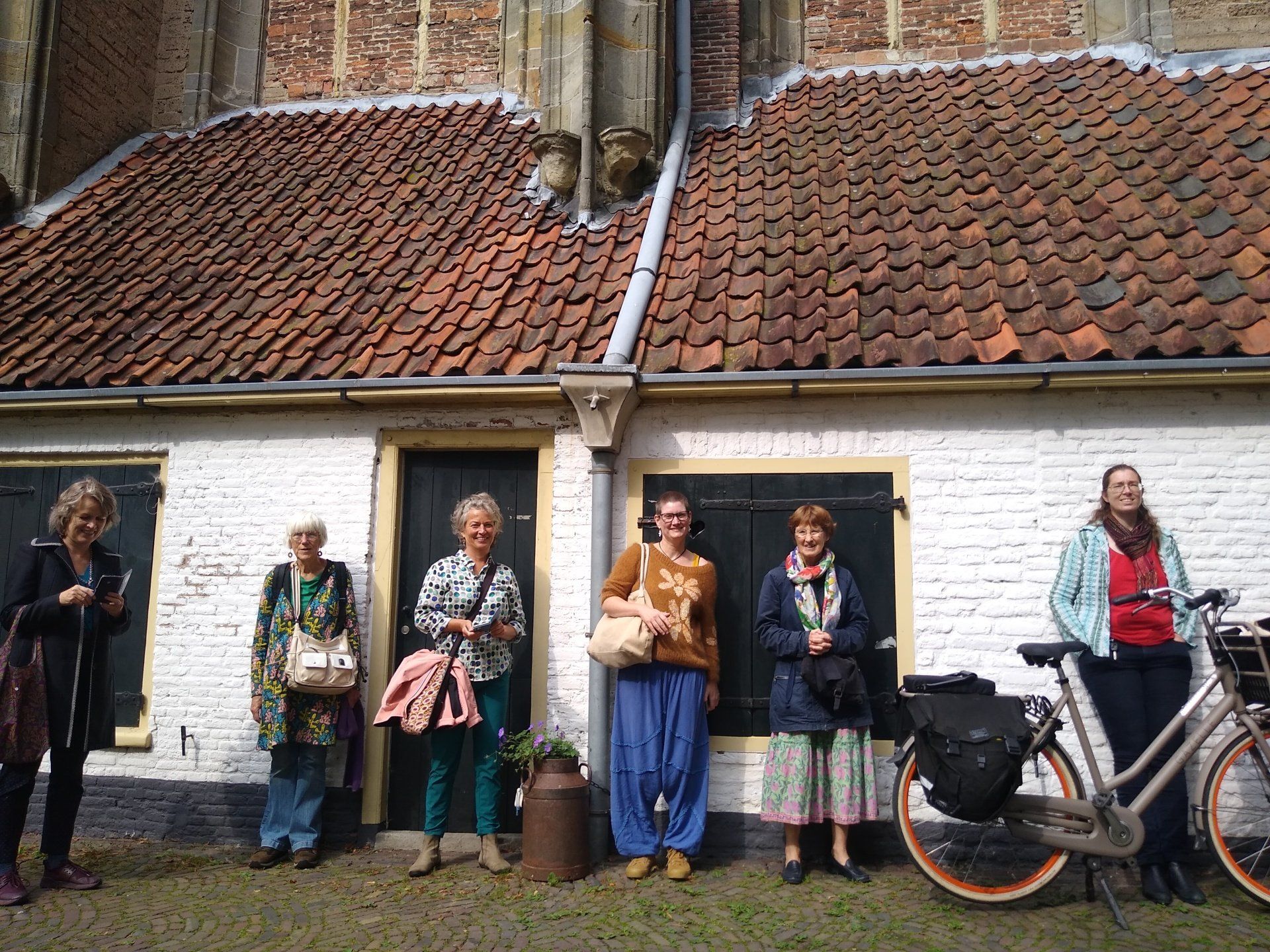 Zes mensen staan in een rij voor een wit, bakstenen huisje met een pannendak, waarvan er één tegen een fiets leunt.