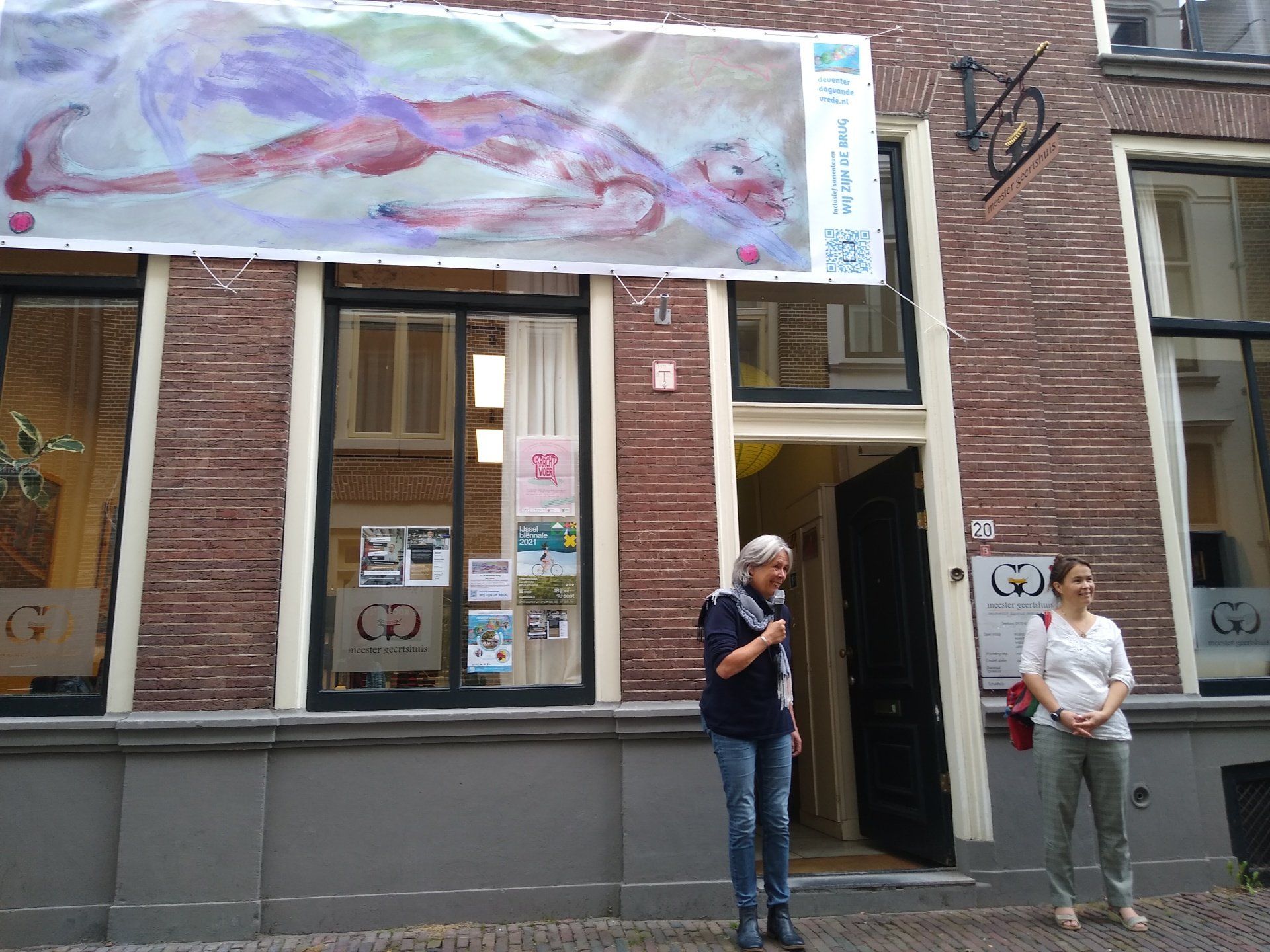 Twee mensen staan buiten een bakstenen gebouw met een groot spandoek boven de ingang waarop een geschilderde figuur is afgebeeld.