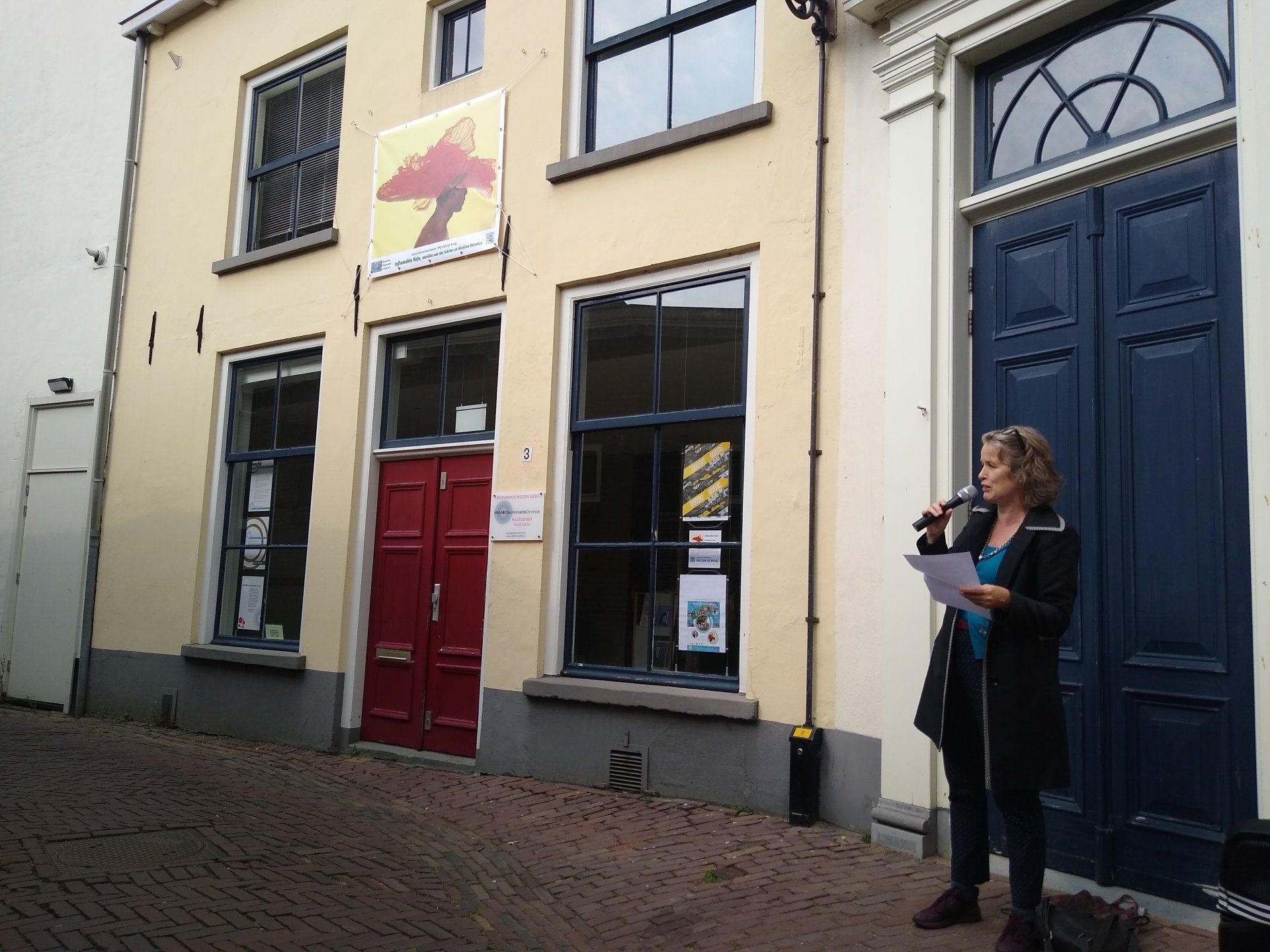Een persoon met een microfoon en aantekeningen staat voor een geel gebouw met een rode deur en geeft een presentatie.
