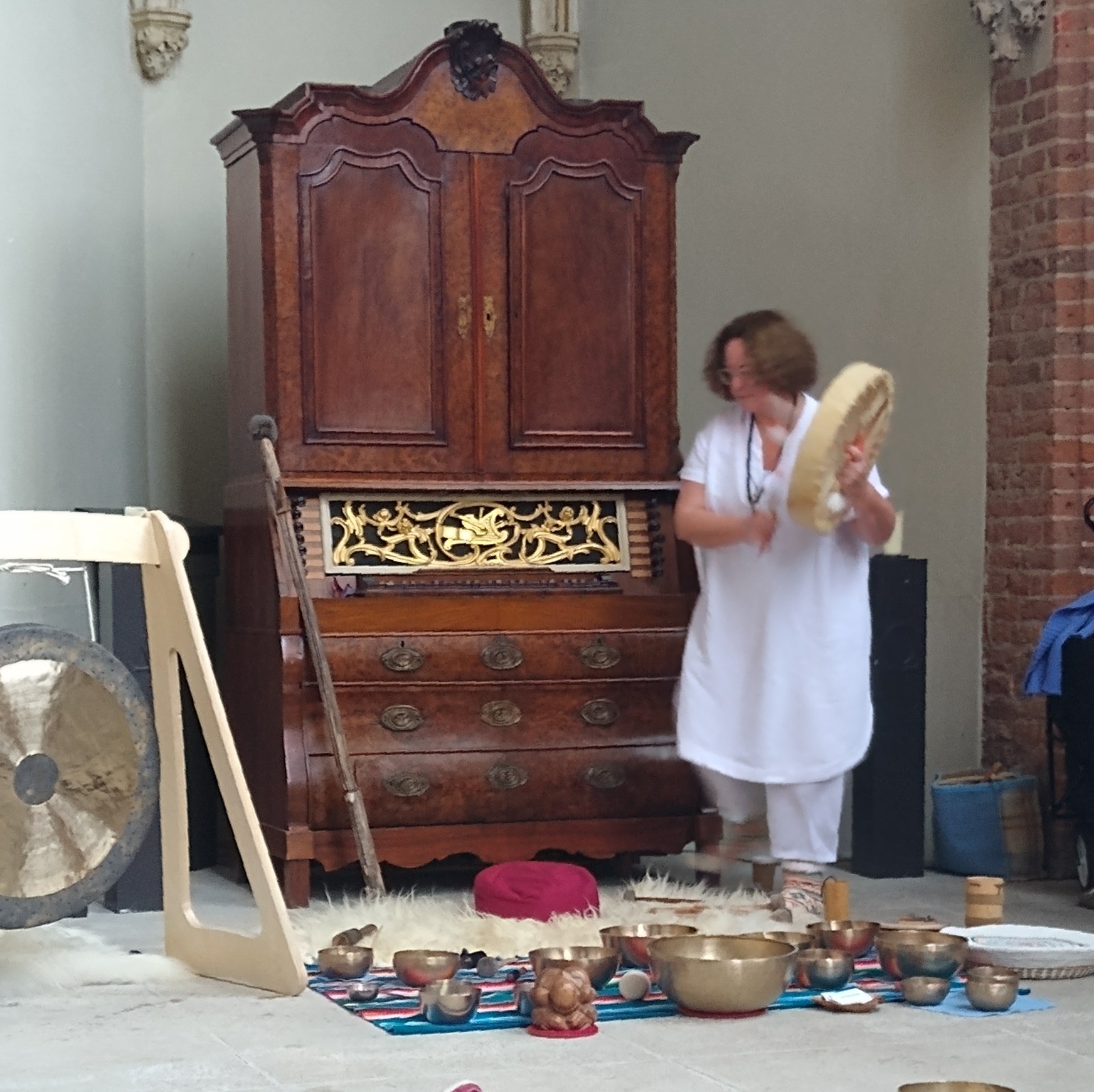 Een persoon in witte kleding houdt een ronde trommel vast voor een hoge houten kast, omringd door klankschalen op een tapijt.