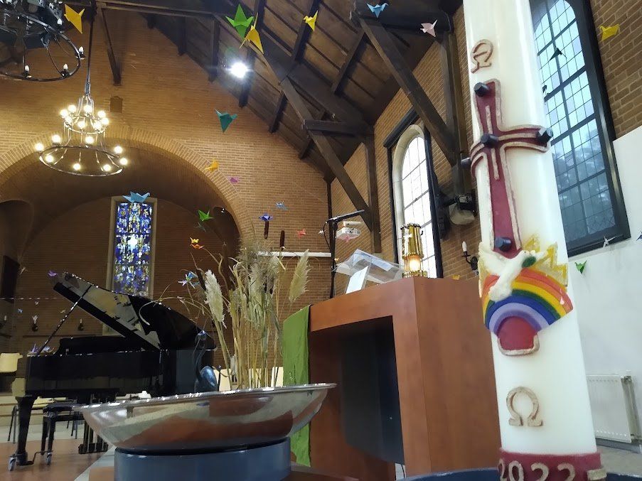 In een kerk staat een grote, versierde paaskaars voor 2022 naast een doopvont, een vleugel en glas-in-loodramen.