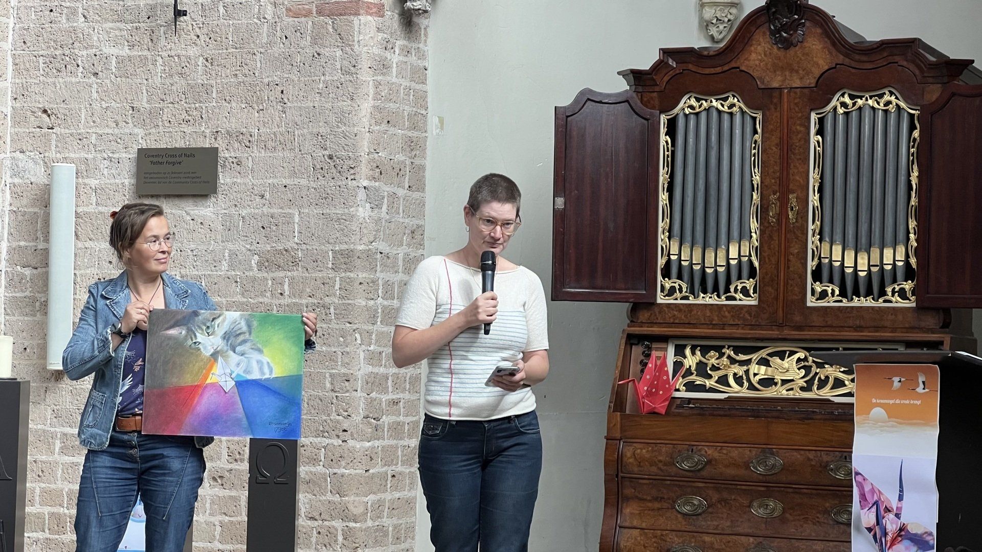 Twee mensen staan in een stenen ruimte vlakbij een sierlijk orgel. De ene houdt een kleurrijk schilderij vast, terwijl de andere in een microfoon spreekt.
