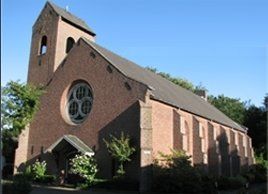 Een kerk van rode baksteen met een hoge klokkentoren, een opvallend rond raam en een zadeldak, afgetekend tegen een helderblauwe hemel.