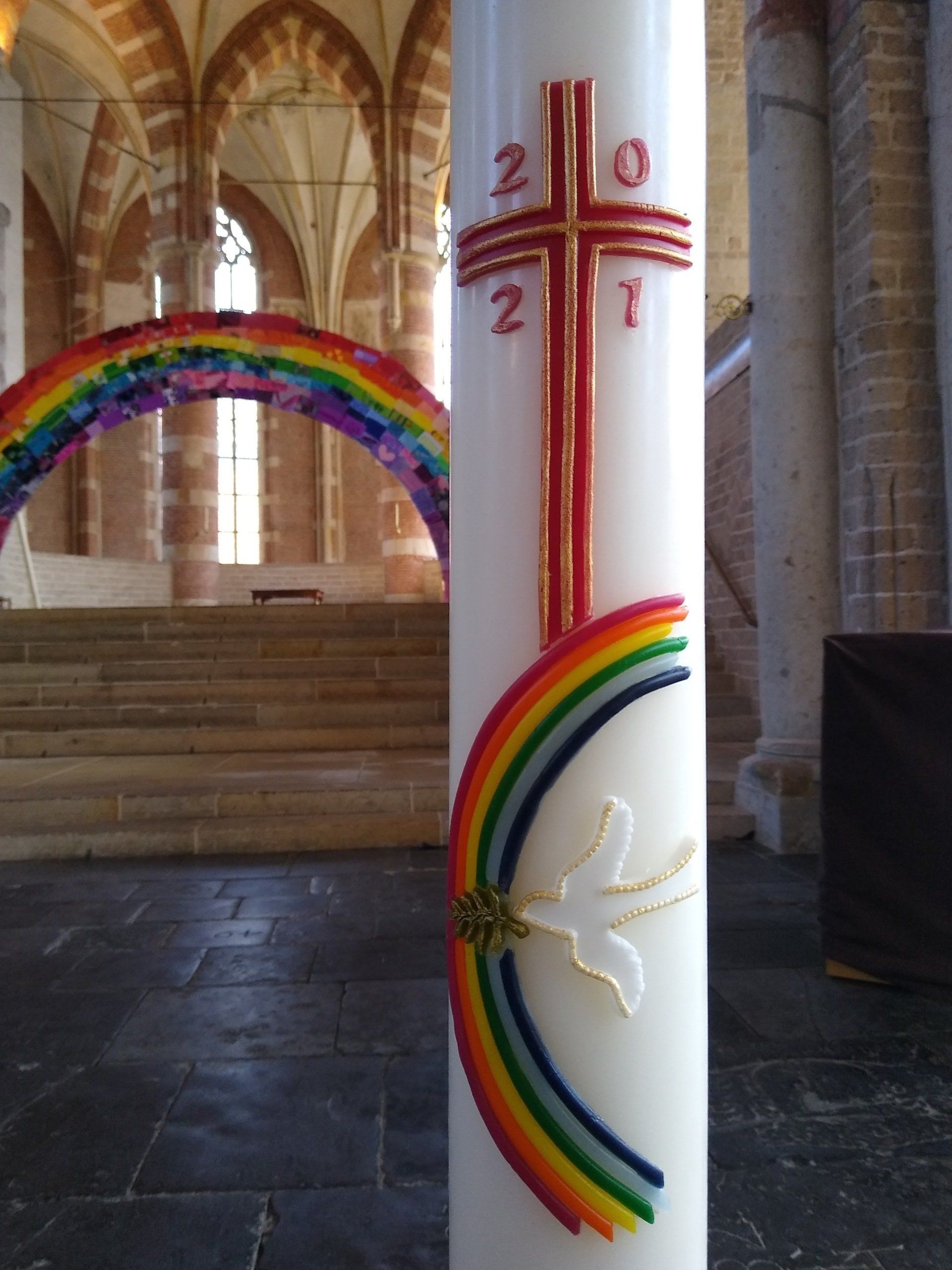 Een paaskaars met een kruis, het jaartal 2021 en een regenboogduif, geplaatst in een kerk met een regenboogboog op de achtergrond.