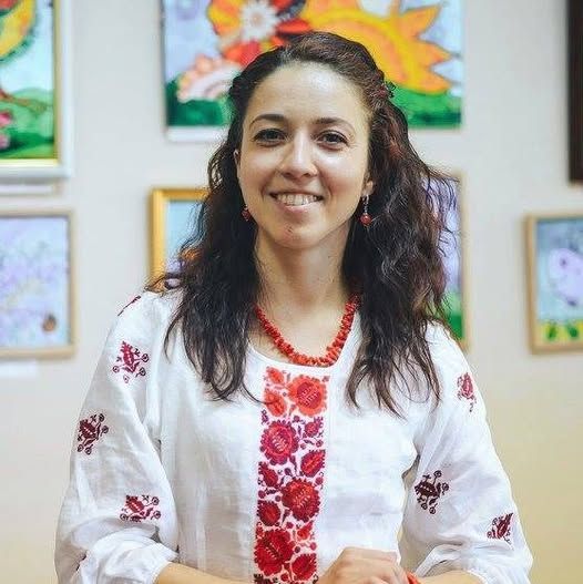 Katrin Poliarush, gekleed in een wit, geborduurd shirt met rode bloemenmotieven, staat voor een ingelijst kunstwerk.