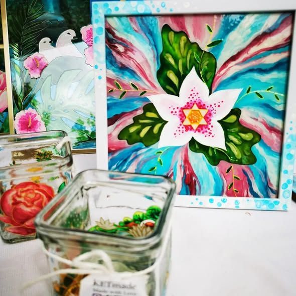 Een vierkant schilderij van een witte bloem in het midden tegen een kleurrijke, abstracte achtergrond, met glazen potten op de voorgrond.
