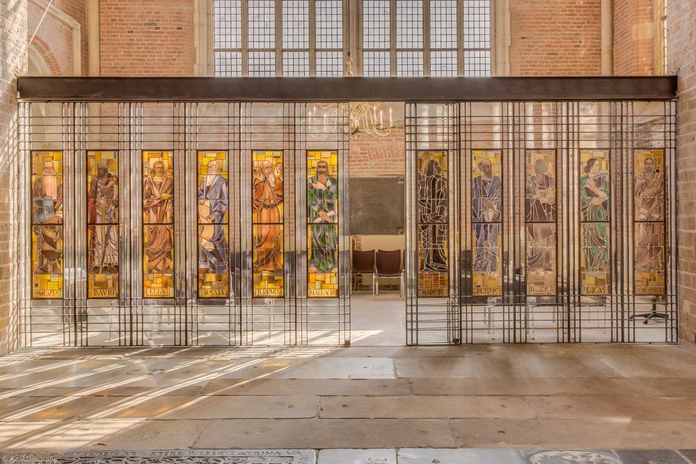 Een decoratief metalen scherm met glas-in-loodpanelen waarop figuren zijn afgebeeld, geplaatst in een zonovergoten stenen hal.