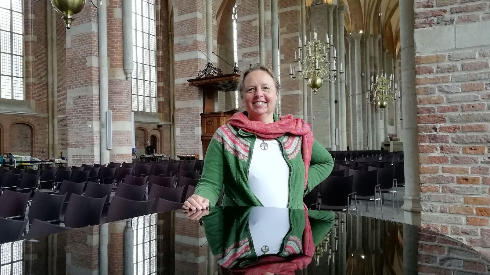 Een glimlachend persoon in een groen vest en een roze sjaal leunt tegen een zwarte piano in een grote, gewelfde bakstenen kerk.