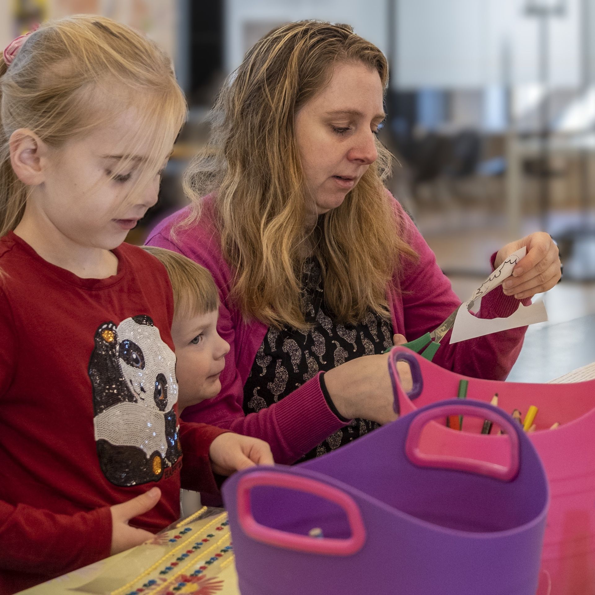 Een volwassene en twee kinderen zitten samen aan een tafel papier te knippen voor een knutselproject, vlakbij kleurrijke opbergbakken.