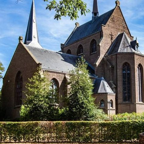 Een hoge bakstenen kerk met een spitse torenspits en boogvensters, omlijst door groene bomen en een haag onder een blauwe hemel.