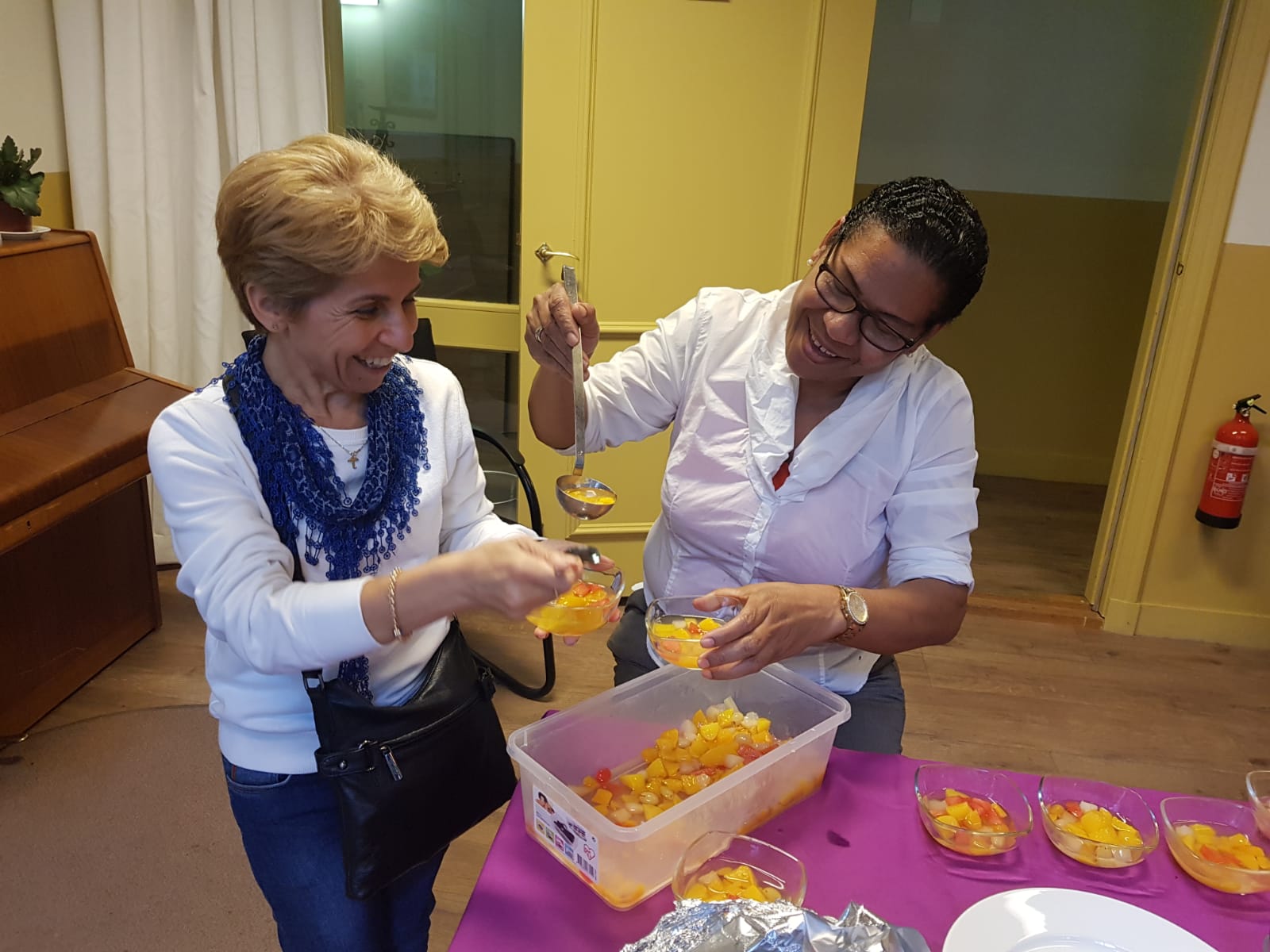Twee mensen glimlachen terwijl ze fruitsalade in kleine plastic schaaltjes serveren op een paars gedekte tafel.