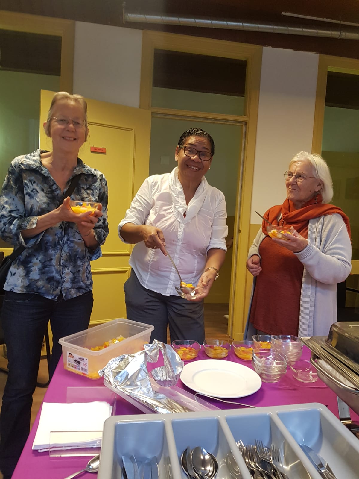 Drie mensen glimlachen terwijl ze kleine porties eten uit bakjes opscheppen en opeten aan een tafel met bestek en borden.