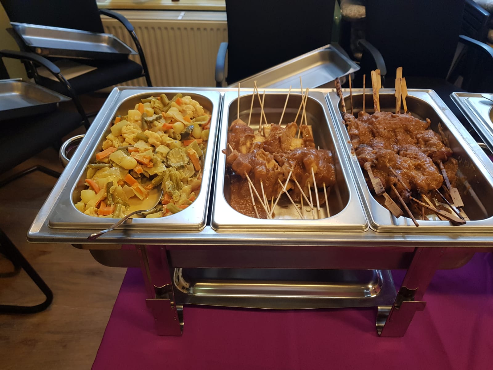 Een metalen warmhoudschaal op een paars tafelkleed, gevuld met gekookte groenten en vleesspiesjes.