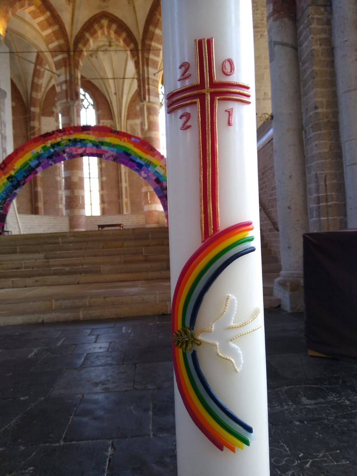 Een paaskaars uit 2021, versierd met een regenboog en een witte duif, staat in een kerk, met een grote regenboogboog op de achtergrond.