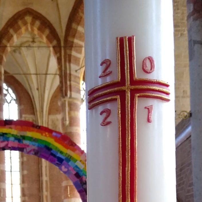 Een close-up van een paaskaars uit 2021 met een rood kruis, met op de achtergrond een wazig beeld van het interieur van een kerk en een regenboog.