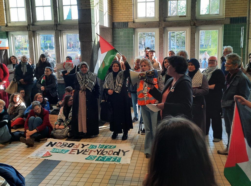Een groep mensen verzamelt zich binnen voor een protest. Ze zwaaien met Palestijnse vlaggen en houden een spandoek vast met de tekst: