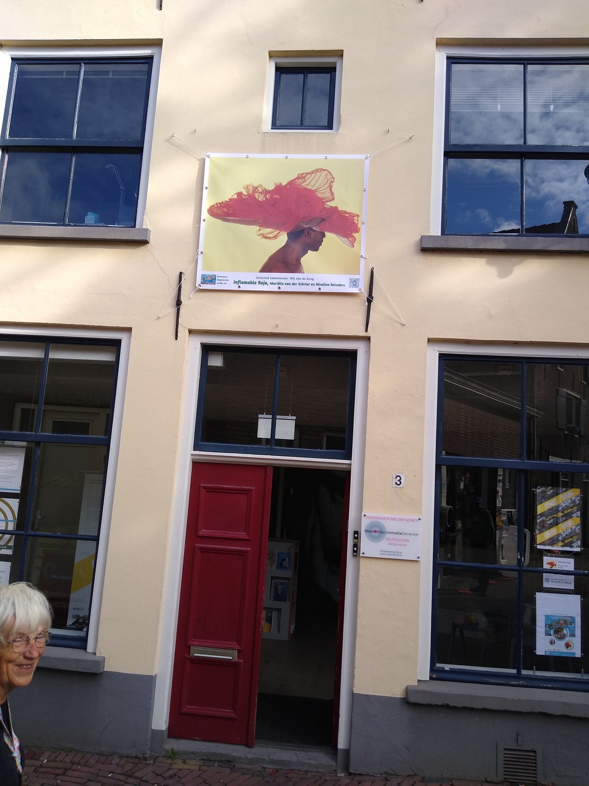 Een lichtgekleurd gebouw met een rode deur, een centrale kunstposter met een figuur in een rode wolk en een persoon links daarvan.