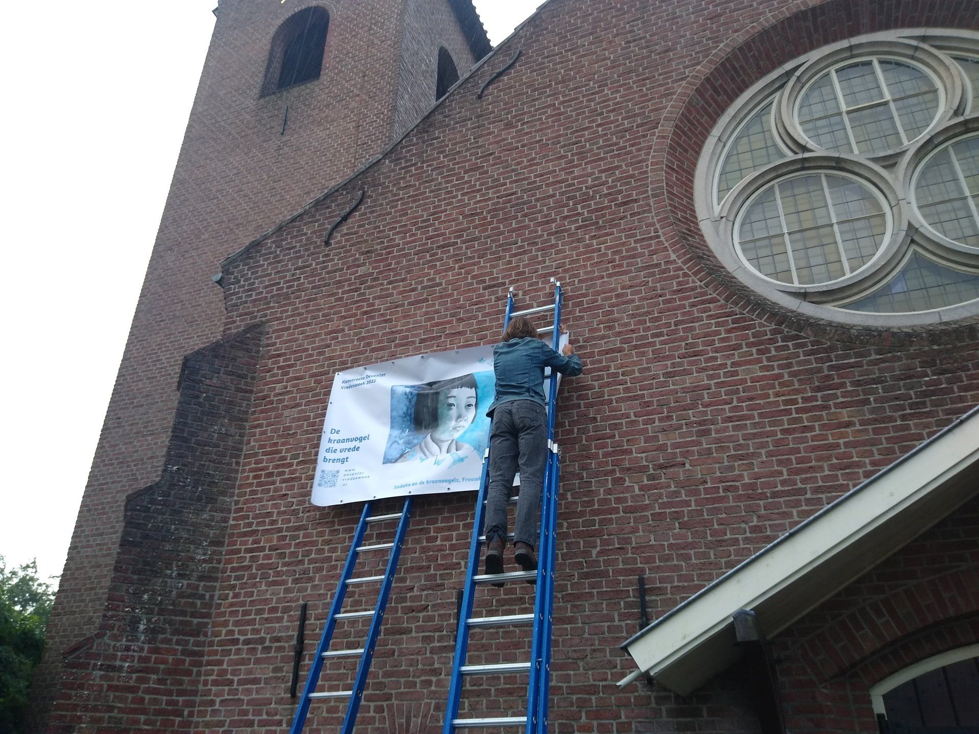 Een persoon op een blauwe ladder bevestigt een wit bord aan de rode bakstenen muur van een kerkgebouw.