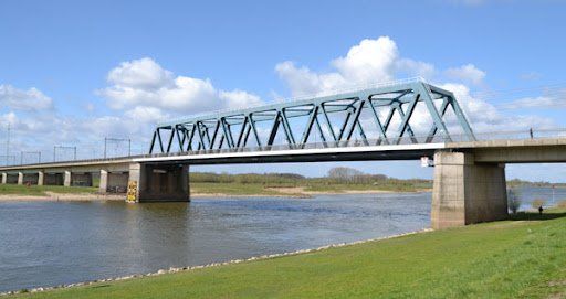 Een blauwe metalen vakwerkbrug overspant een brede rivier onder een blauwe, bewolkte hemel, met groene, met gras begroeide oevers op de voorgrond.
