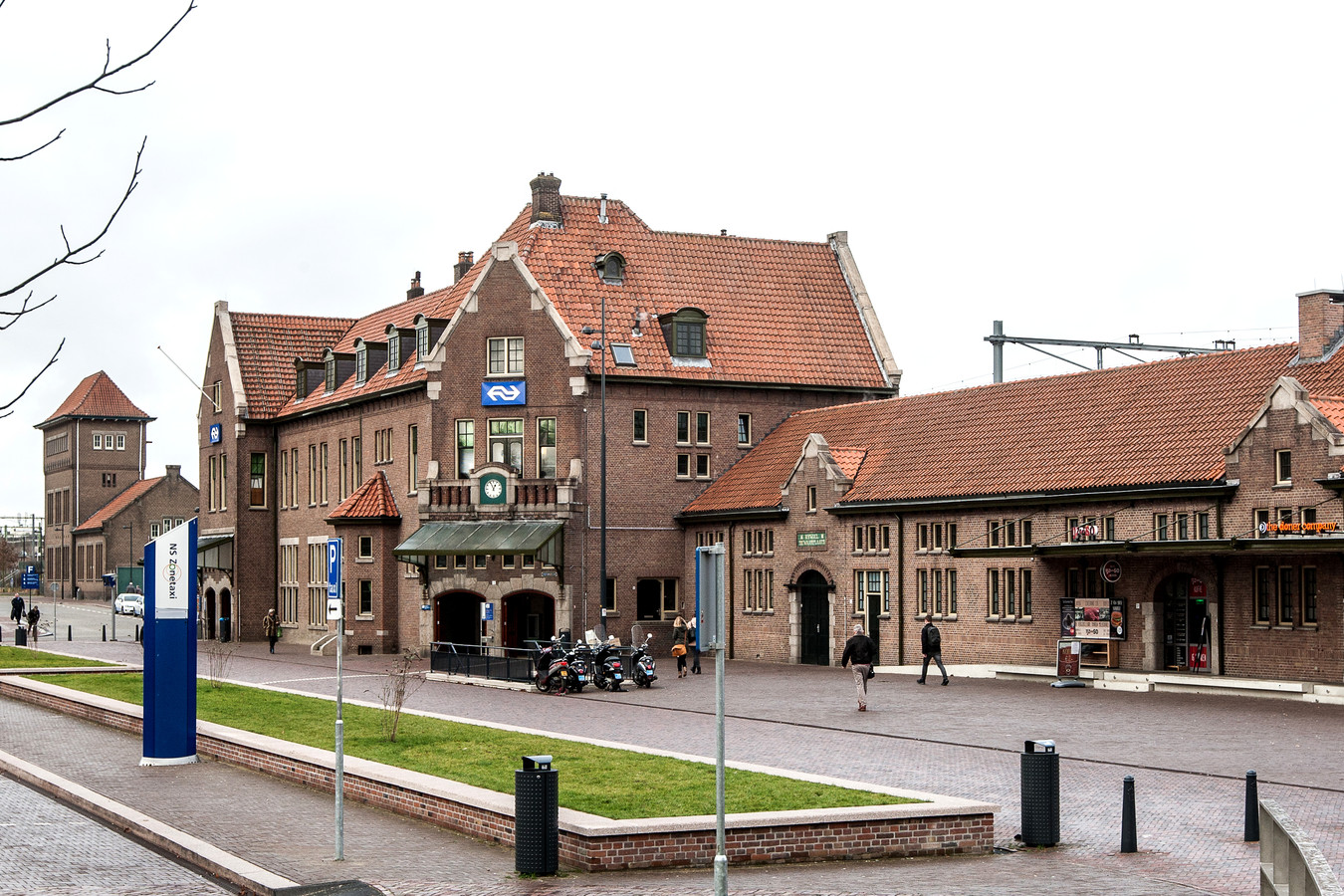 Het historische bakstenen station in Winterswijk, Nederland, met een pannendak, parkeerplaats en blauwe bewegwijzering.