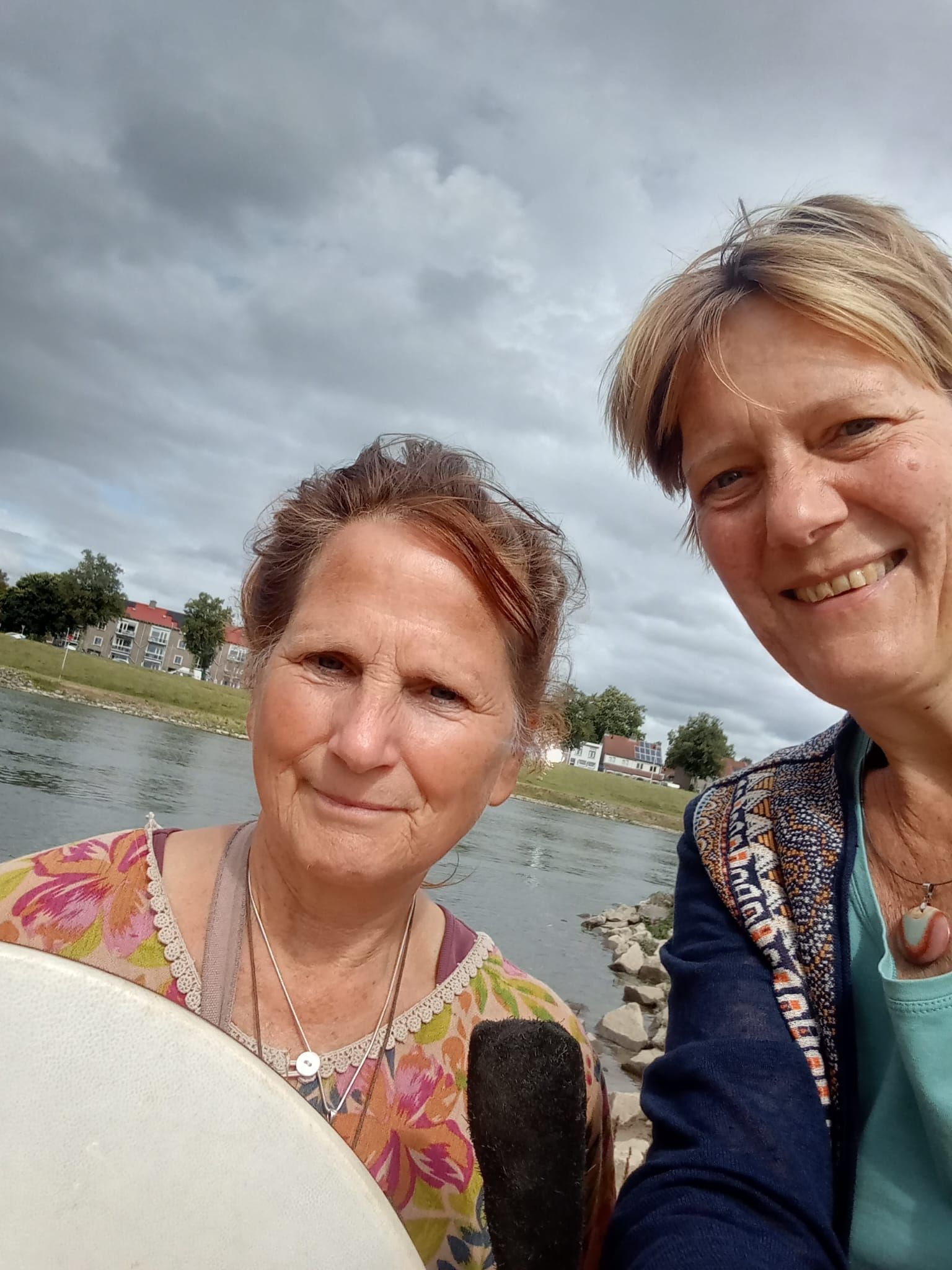 Twee lachende personen maken een selfie in de buitenlucht, vlakbij een rivieroever, onder een bewolkte hemel.