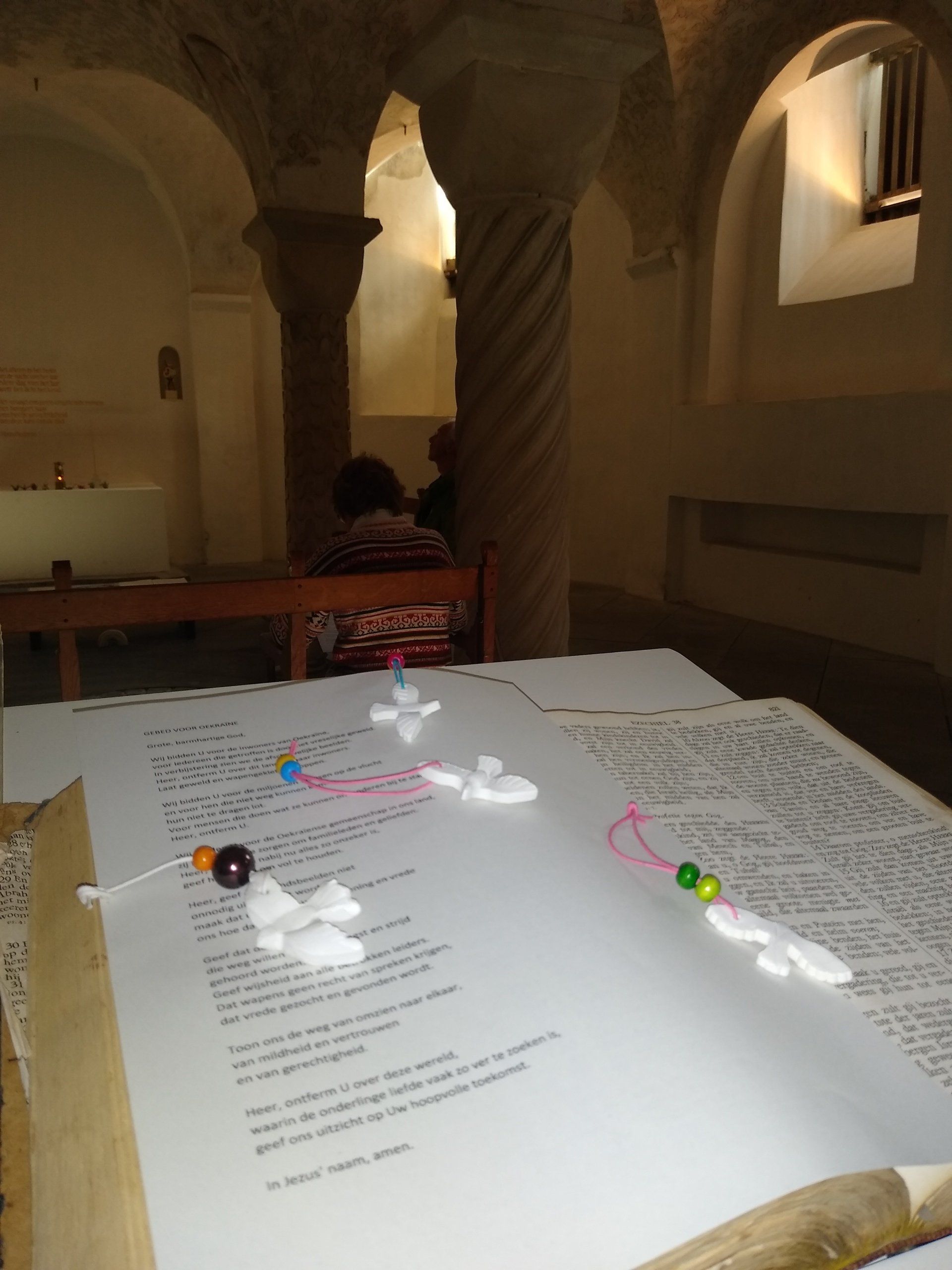 Een gebedenboek met kleine kralenkruisjes staat op een houten standaard in een schemerig verlichte stenen kerk met gewelfde pilaren.