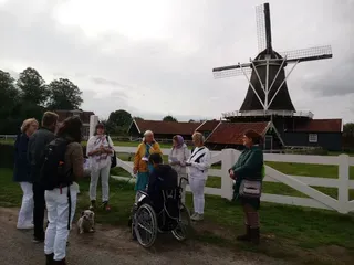 Een reisgezelschap staat buiten bij een wit hek, met op de achtergrond een grote, traditionele Nederlandse windmolen.