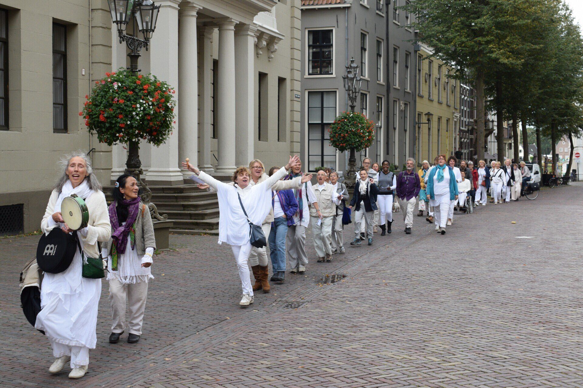 Een groep mensen in witte kleding loopt en viert feest op een geplaveide straat voor een gebouw met stenen pilaren.