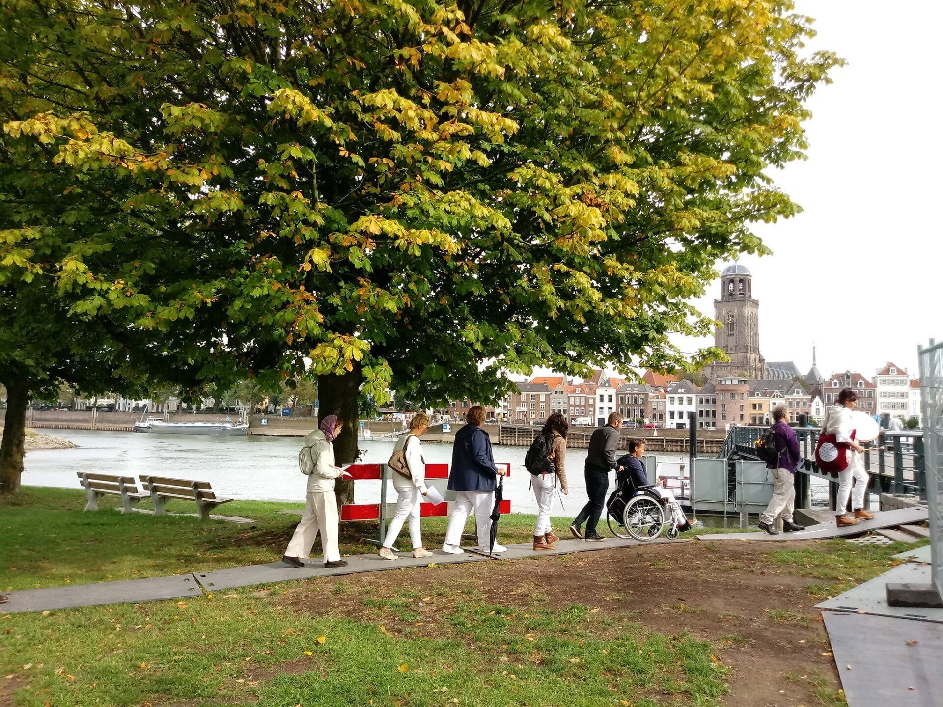 Een groep mensen, waaronder iemand in een rolstoel, loopt langs een pad aan de rivier onder een grote herfstboom.