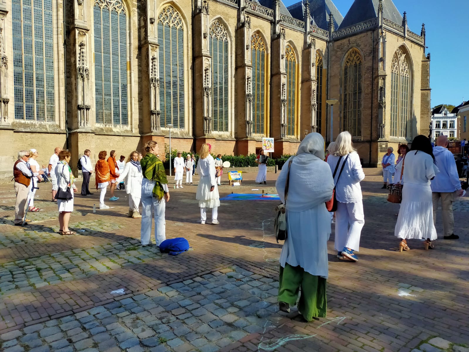 Op een zonnige dag staan mensen in lichtgekleurde kleding op een plein voor een grote, gotische stenen kerk.