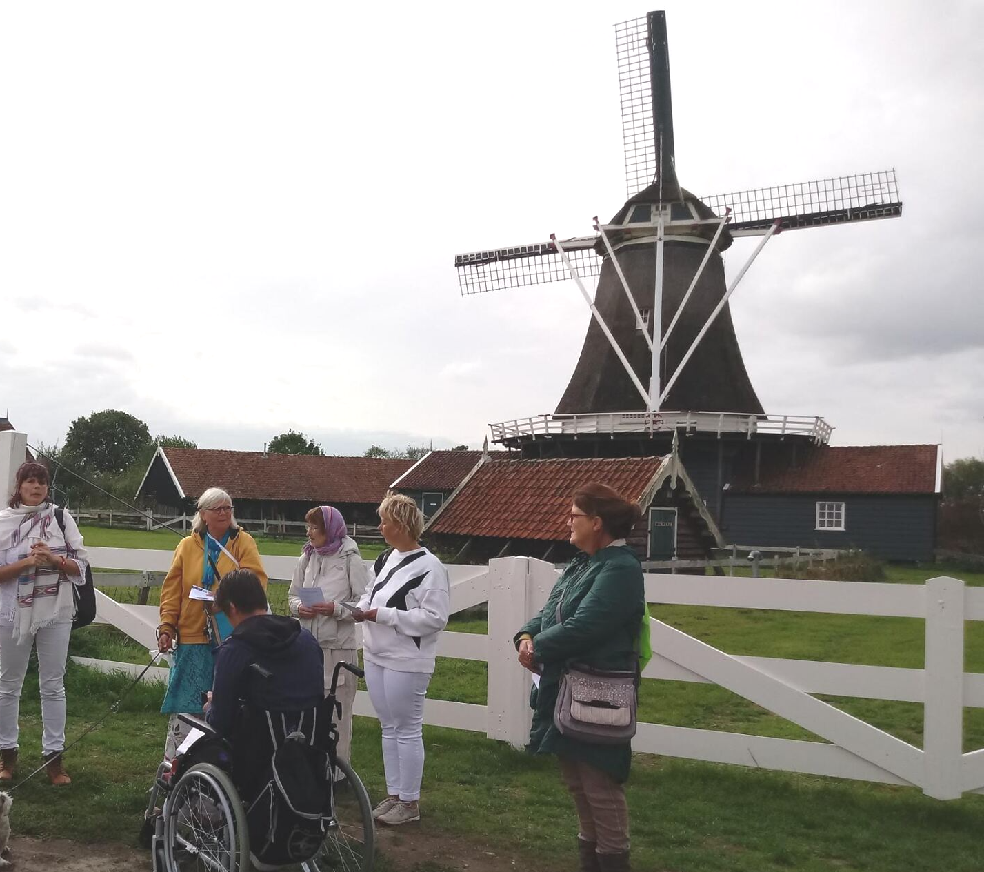 Een groep mensen, onder wie iemand in een rolstoel, staat buiten een hoge, donkere houten windmolen op een grasveld.