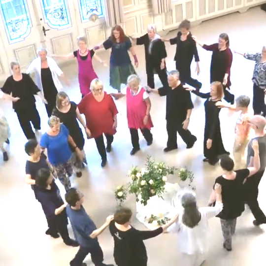 Een diverse groep mensen staat in een grote kring in een lichte ruimte, hand in hand rond een centraal bloemstuk.