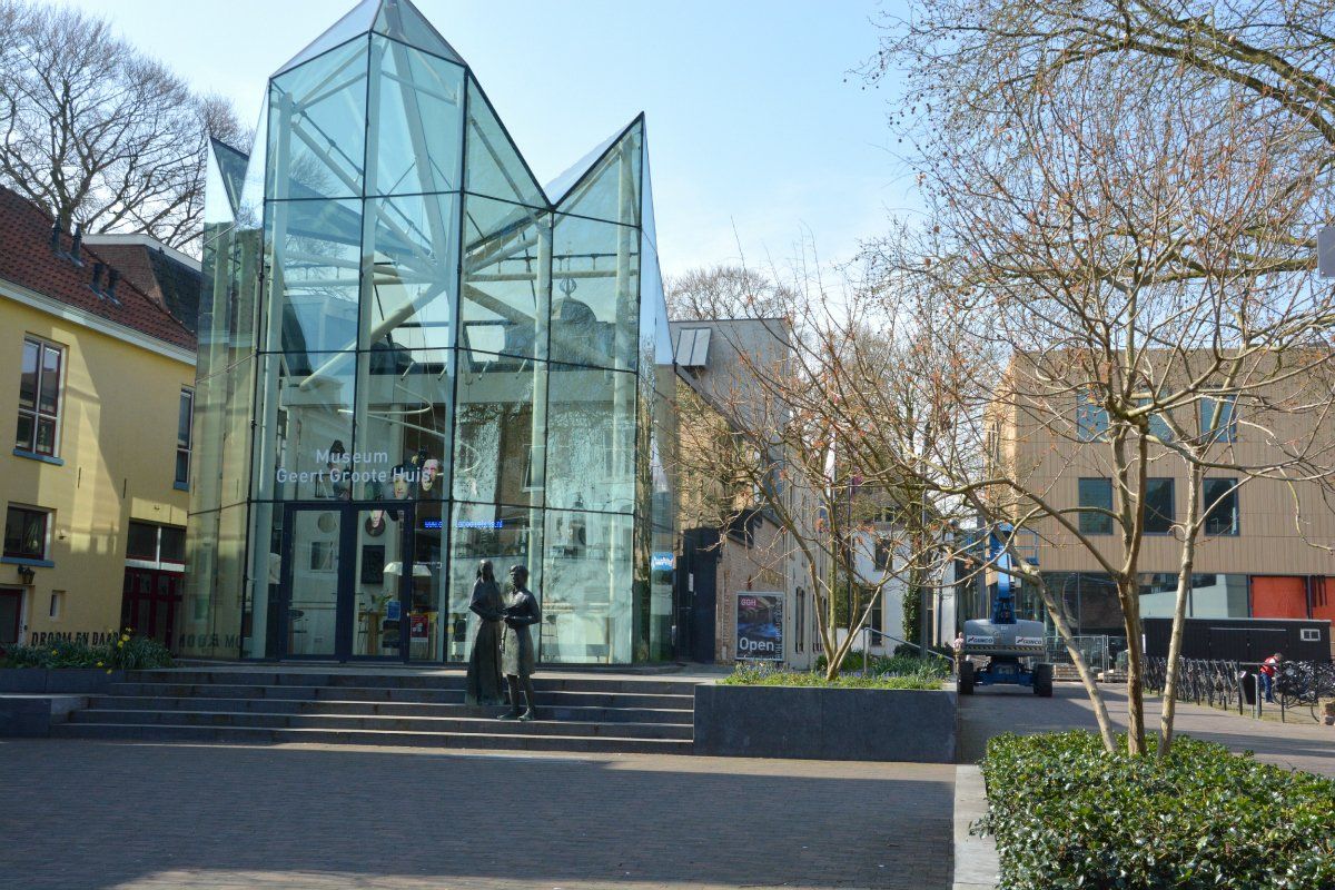 Modern glazen gebouw met een puntig dak, omgeven door bomen en historische architectuur op een openluchtplein.