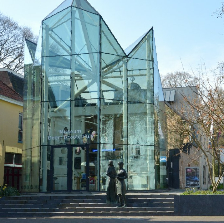 Het museum Geert Groote Huis is gevestigd in een glazen gebouw met twee beelden ervoor, aan een trap.