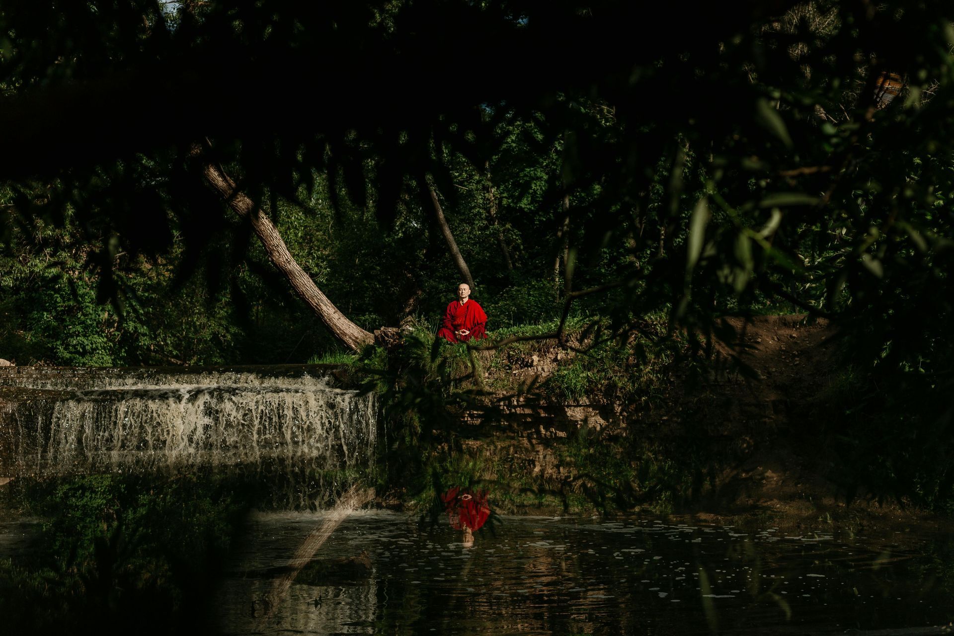 Een persoon in een felrood gewaad zit meditatief op de oever van een beekje, vlakbij een kleine, rotsachtige waterval.