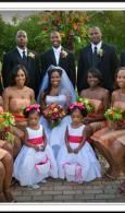 Formal wedding party posing outdoors, with two women in white dresses and floral bouquets seated in front.
