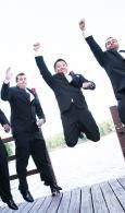 Three businesspeople in suits jumping with raised fists outdoors on a wooden deck
