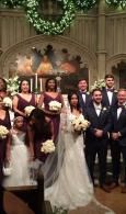 Wedding party posing indoors in front of a decorated altar, with the bride in white holding a bouquet.