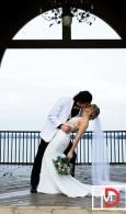 Couple in wedding attire kissing by a seaside railing under an arched structure