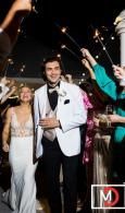 Man in white tuxedo holding a drink at a lively nighttime event, surrounded by cheering guests