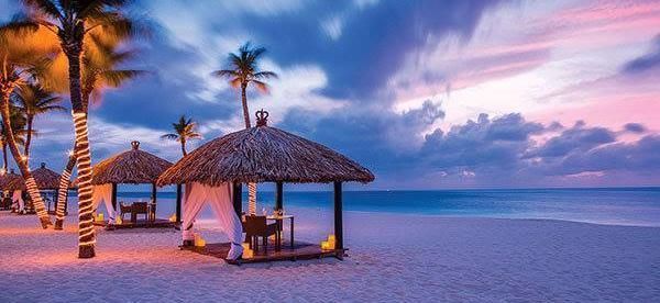 Tropical beach at sunset with thatched cabanas, palm trees, and calm blue water under pink clouds