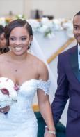 Bride in a white dress holding a bouquet, smiling beside a man in a dark suit outdoors.