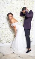 Bride in white gown posing beside groom in dark suit against a white floral backdrop