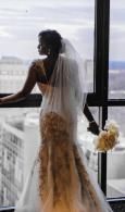 Bride in a lace wedding gown and veil standing by a window with a city view and bouquet beside her