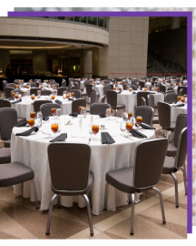 Round banquet tables with white tablecloths and gray chairs set in a large indoor venue.
