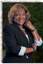 Smiling woman in a dark blazer posing with hand on chin against a blurred green background
