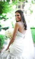 Bride in a white wedding dress and veil, smiling outdoors with a green garden background