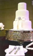 White tiered wedding cake on a decorative silver stand with silver floral accents and forks on a napkin nearby