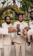 Three men in beige suits stand outdoors, smiling and holding a bottle of champagne.
